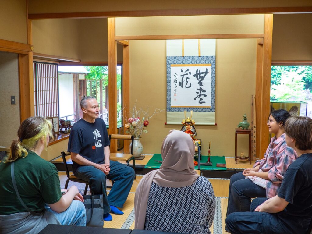 Inside the study hall of Tokoji Temple, iCLA students and Professor Reed are sitting in a group and discussing Zen concepts of mindfulness. The room is in Japanese style with tatami mats, and tastefully decorated with a hanging scroll, a mini set of samurai armor, and other Japanese objects.