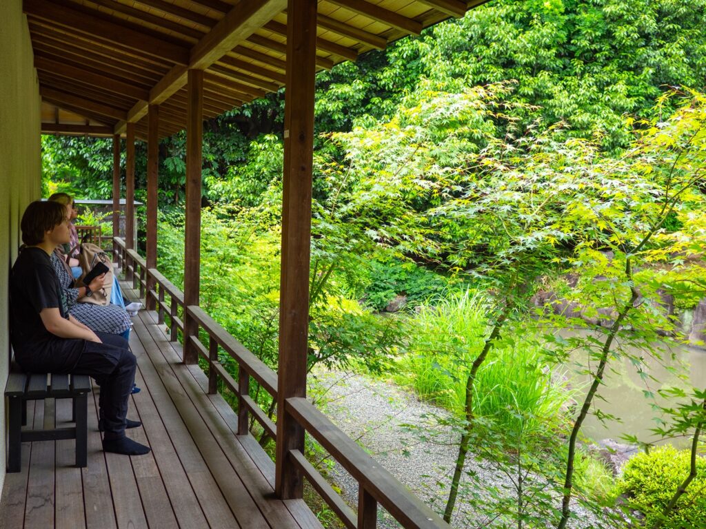iCLA students sit on a bench overlooking a beautiful garden. The garden has a pond, and the greenery is lush.