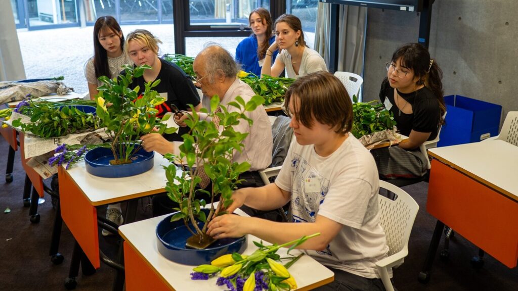 iCLA students sit at desks in a classroom, learning how to do Japanese flower arrangement or ikebana from a master, who is demonstrating how to arrange the flowers.