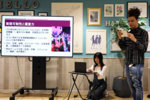 A male iCLA student gives a presentation for the YGU Student Challenge Program. A professor is beside him on a laptop, and there is a large screen showing the presentation.