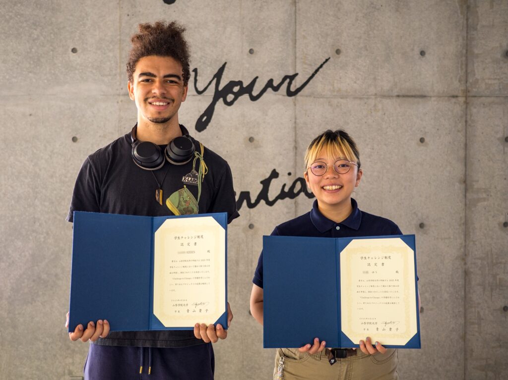 Two smiling iCLA students hold their certificates for the Yamanashi Gakuin University Student Challenge Program, looking proud to be chosen.