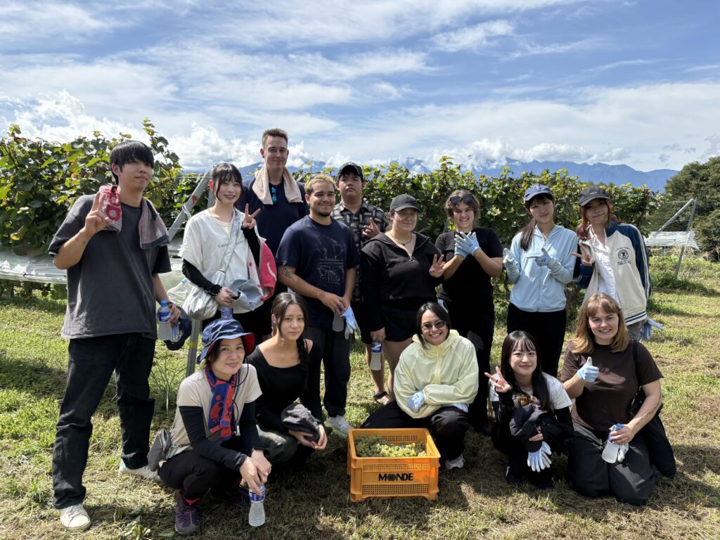 A group of iCLA and Faculty of Business Administration students smile and take a photo together after finishing the workshop to harvest grapes for Yamanashi wine. In front is a Monde Winery basket filled with grapes.