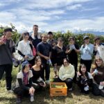 A group of iCLA and Faculty of Business Administration students smile and take a photo together after finishing the workshop to harvest grapes for Yamanashi wine. In front is a Monde Winery basket filled with grapes.