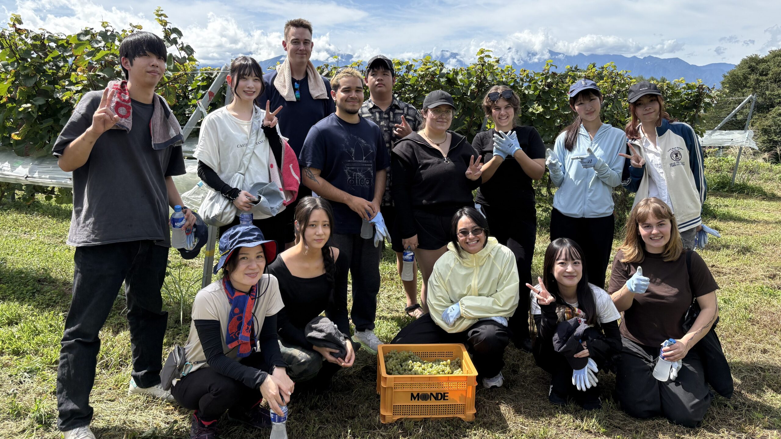 A group of iCLA and Faculty of Business Administration students smile and take a photo together after finishing the workshop to harvest grapes for Yamanashi wine. In front is a Monde Winery basket filled with grapes.