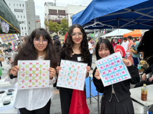 Two iCLA students and a Seishu high school student are smiling and holding up their colorful washi dyeing projects at Global Festa in Kofu.